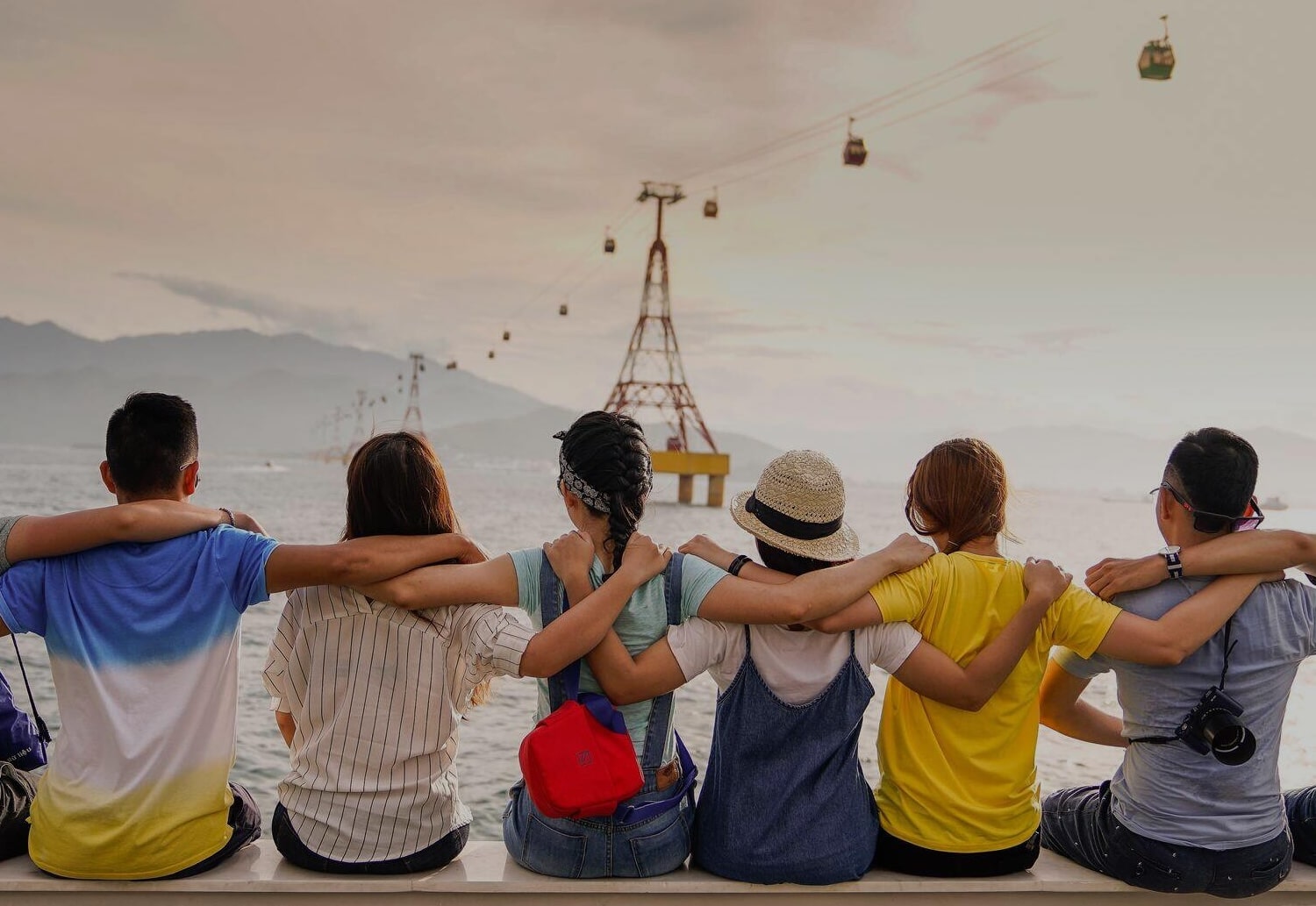 Six friends sit with arms around each other, facing the water as cable cars glide by over a sunset harbor.